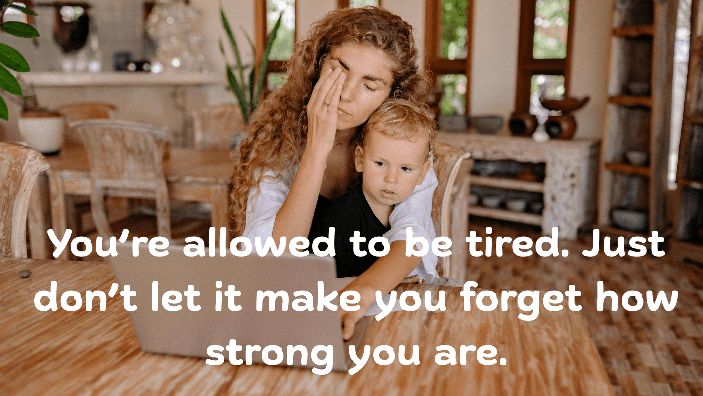 Tired woman working from home while holding a young child on her lap, sitting at a wooden table with a laptop in a cozy room. The text is saying that you have permission to feel tired.